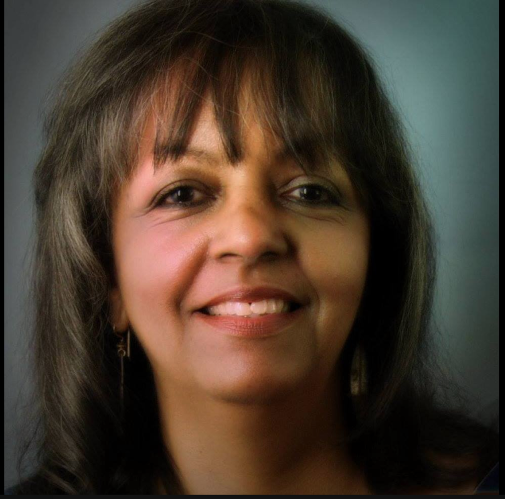 Head shot image of a Black woman in her early 60's smiling into the camera, she has shoulder length hair