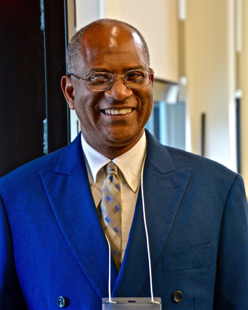 A Back man in his early 60's, wearing a blue suit, tie and glasses, smiles into the camera
