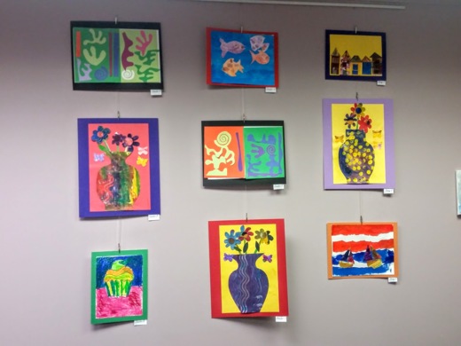 Multiple art works from students hung on a wall. Some are drawings of vases with flowers another is a cupcake 