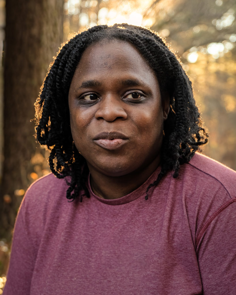 A Black woman in her 30's looking off into the distance with the woods and the sunset behind her, hair is black shoulder length, she is wearing a red shirt