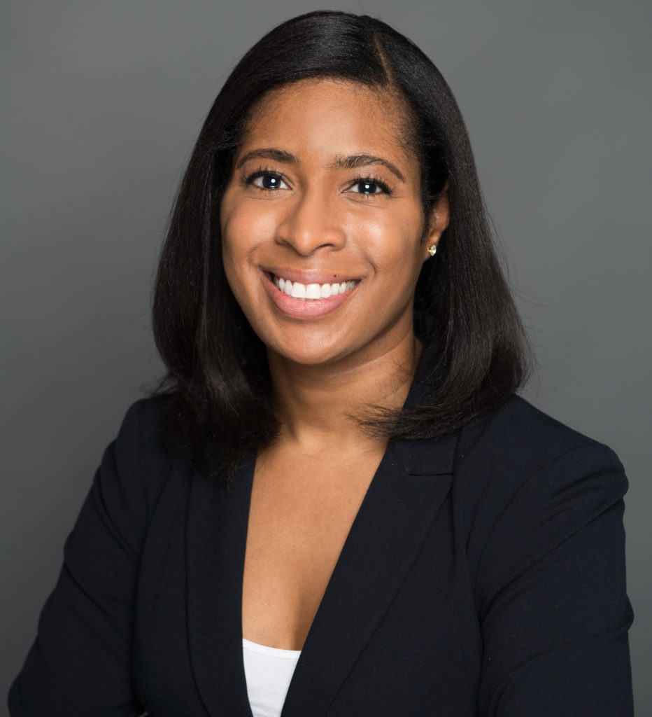 A Black woman in her 30's with shoulder length hair in a black suit is smiling into the camera