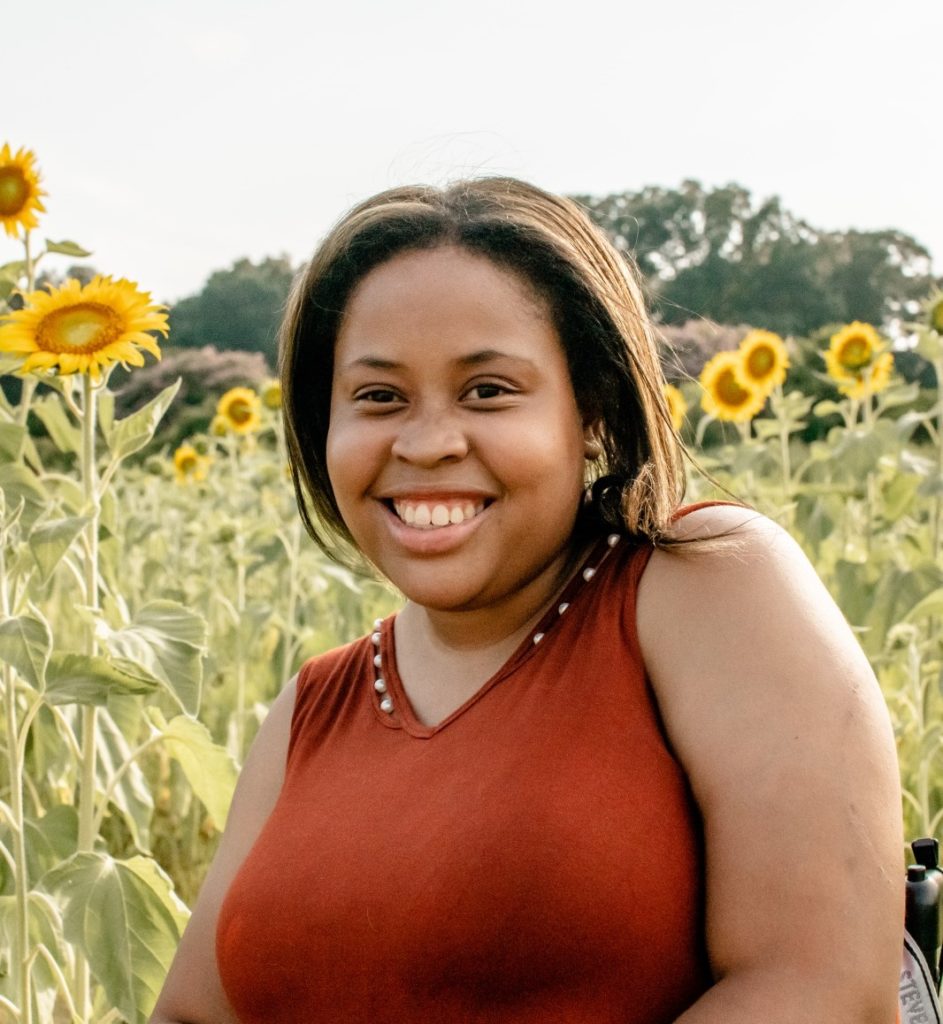 A Black woman in her 20's sitting with a field of sunflowers behind her, she has on a v neck copper shirt