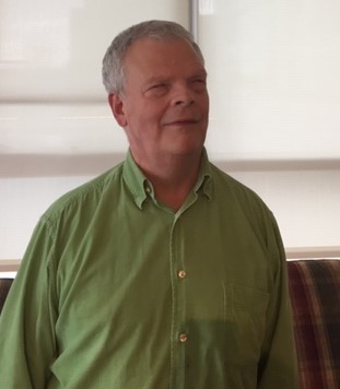 A white man in his 60's with a green button up shirt is smiling into the camera
