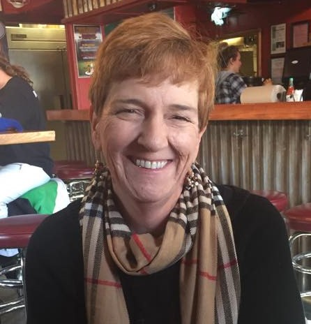 A white woman in her early 60's with short red hair and a brown striped scarf is smiling into the camera