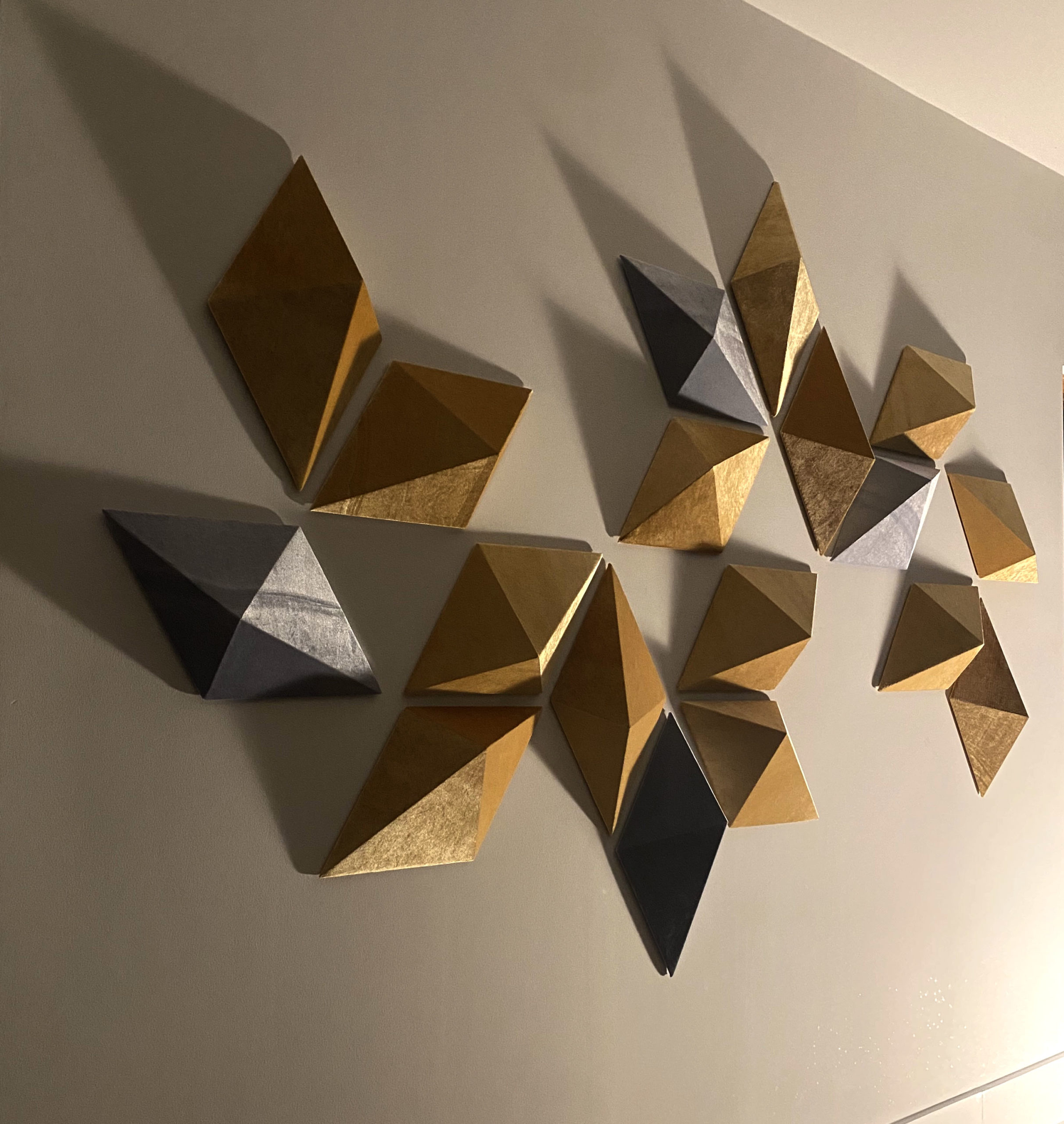 Multiple 3D Dimond shapes of gold and silver are hung on a wall with a light off to the side causing shadows to cast to the left of them.