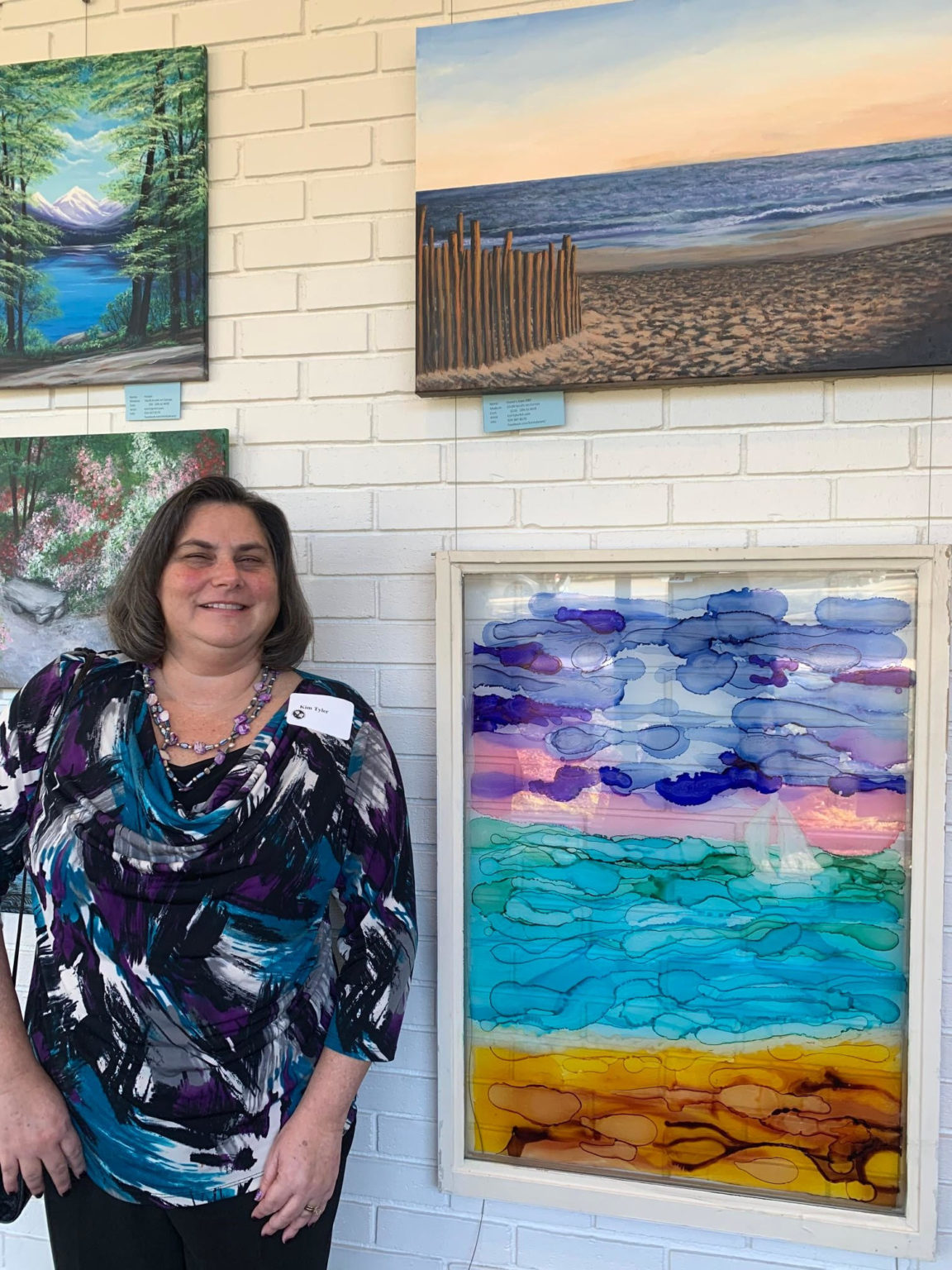Kim Tyler stands next to a large abstracted landscape ink painting on a windowpane. Kim is a white woman with shoulder length black hair wearing a blue and purple blouse. Also in view are Kim's acrylic landscape paintings on canvas - a sandy path to the beach and a lake surrounded by trees and mountains.