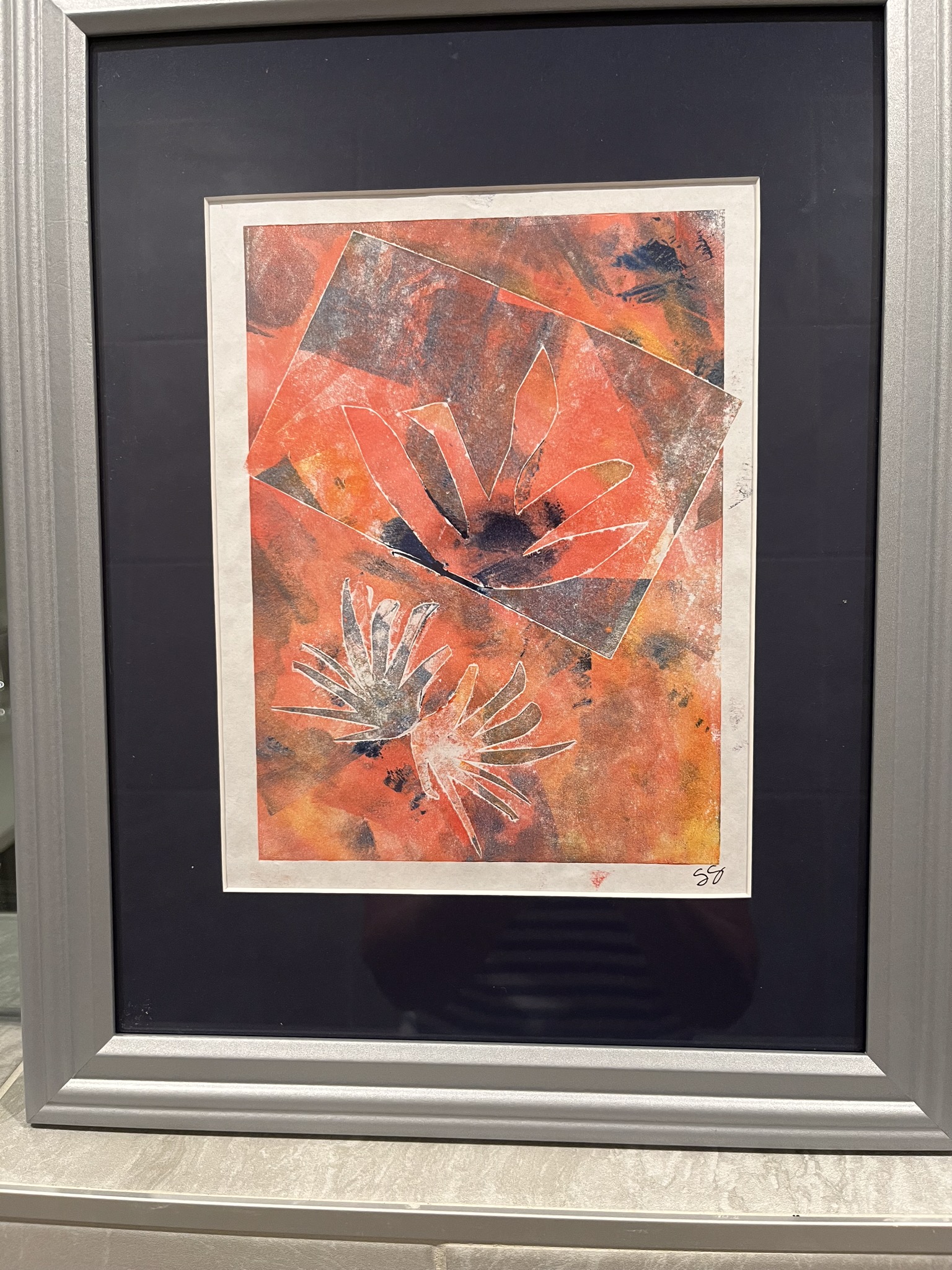 A framed monoprint. The monoprint is many shades of orange and appears to have leaves scattered about. 