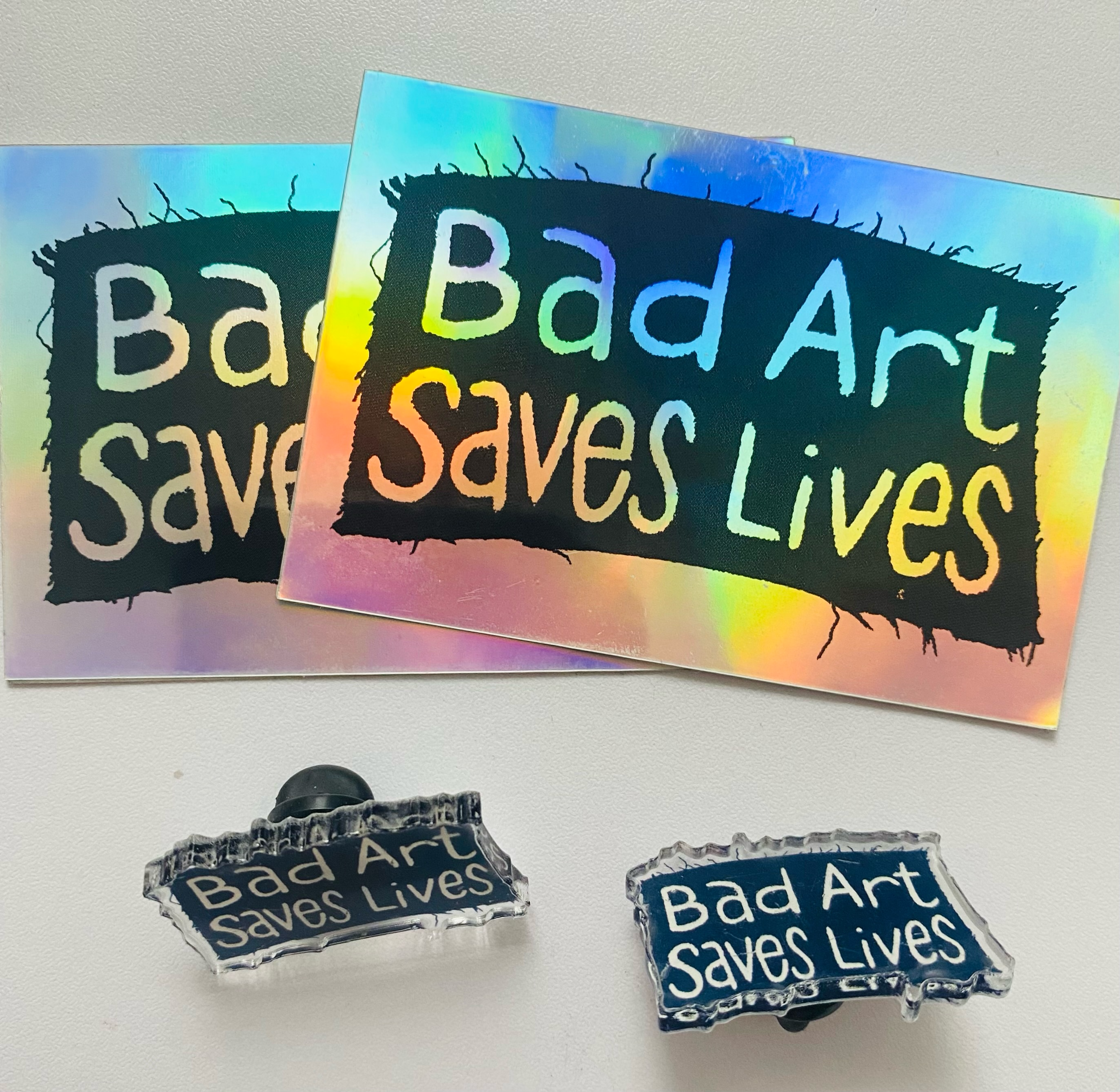 A black patch with "Bad Art Saves Lives" written in white acrylic paint centered in the middle. A holographic sticker with "Bad Art Saves Lives" written in the center, surrounded by a black box. Clear pins with the same design.