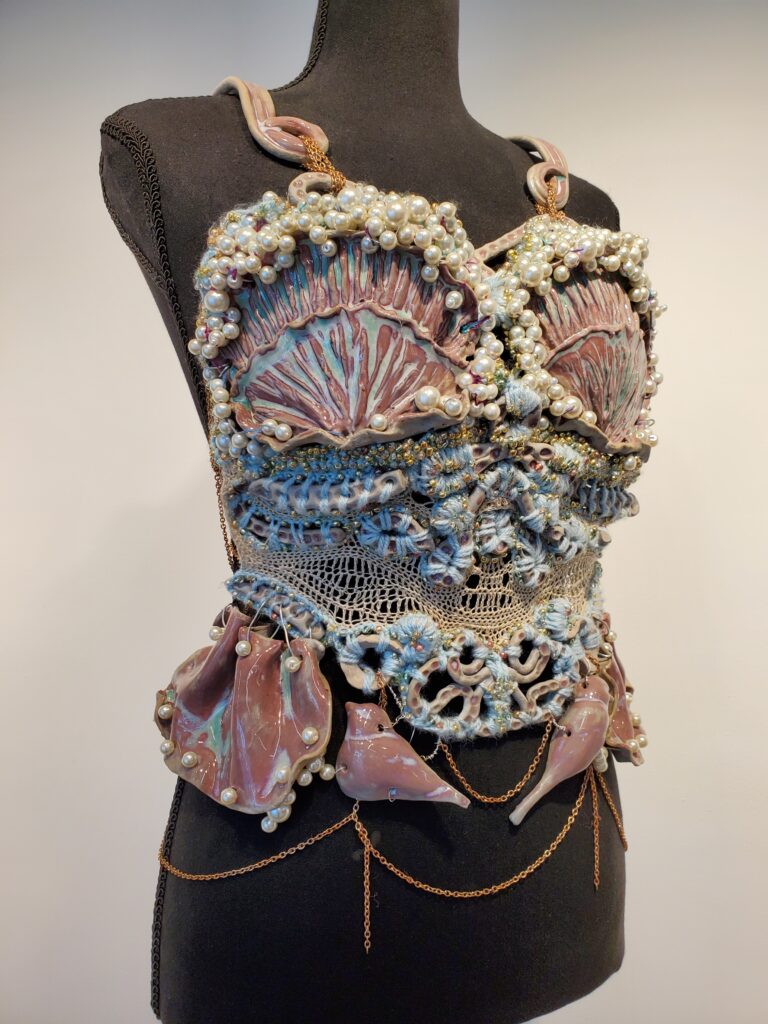 A corset-like top on a black mannequin. The piece is made with a pink and teal ceramic bra and straps, joined together with gold chain. The "bra" piece has pearly beads along it's edges, giving a bubbly or sea foam-like appearance. Matching ceramic pieces in the shapes of swirls make up the middle section of the piece, and are joined with crocheted blue yarn and beige thread. The bottom of the bodice has symmetrical ceramic ruffles and birds on each side.