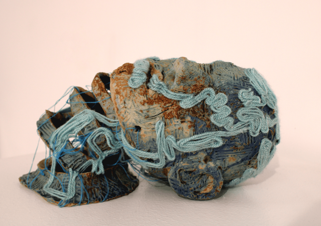 A blue sculpture of a head and neck with a rough texture. The neck is sliced into 4 pieces, which are joined with blue thread. The head is not supported with the loosely connected neck pieces, so it lies on the table. Blue yarn travels across the piece in squiggling lines.
