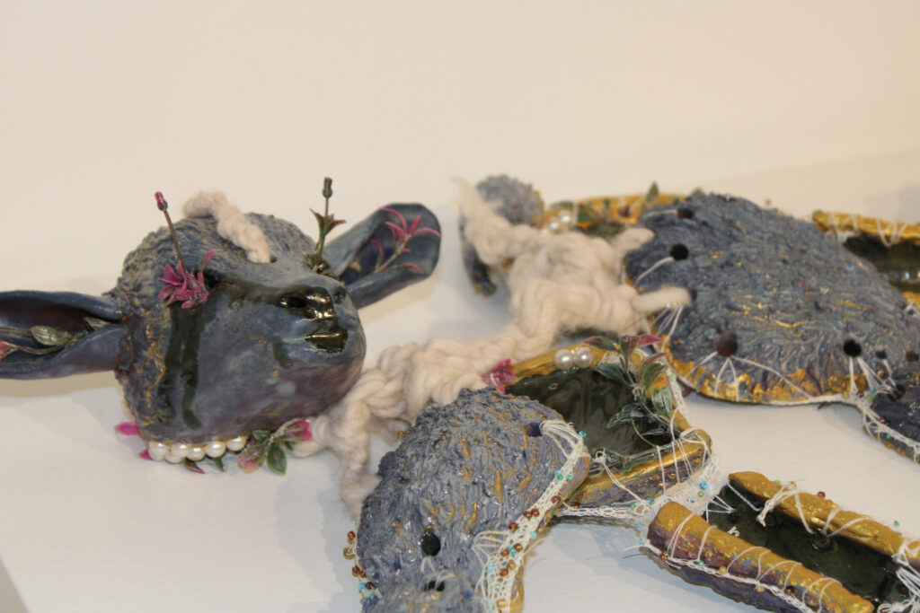 A close-up of a purplish grey lamb sculpture. The lambs limbs are all separate ceramic pieces joined together with crocheted sewing thread strung with small glass beads. The spine is created with thick wool yarn. Faux flowers are growing out of its eye sockets, and the inner edges of the ceramic pieces are lined with gold.