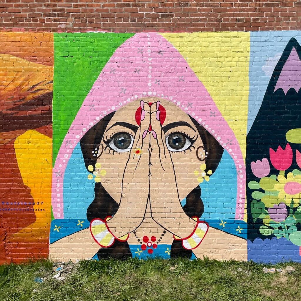 A colorful mural painted on a red brick wall. The painting is of a woman with her hands palms together held up in front of her face. She has light skin and brown hair done in braids. She wears a pink and blue hood on her head and wears a lot of decorative jewelry. On her forehead is a red bindi. 