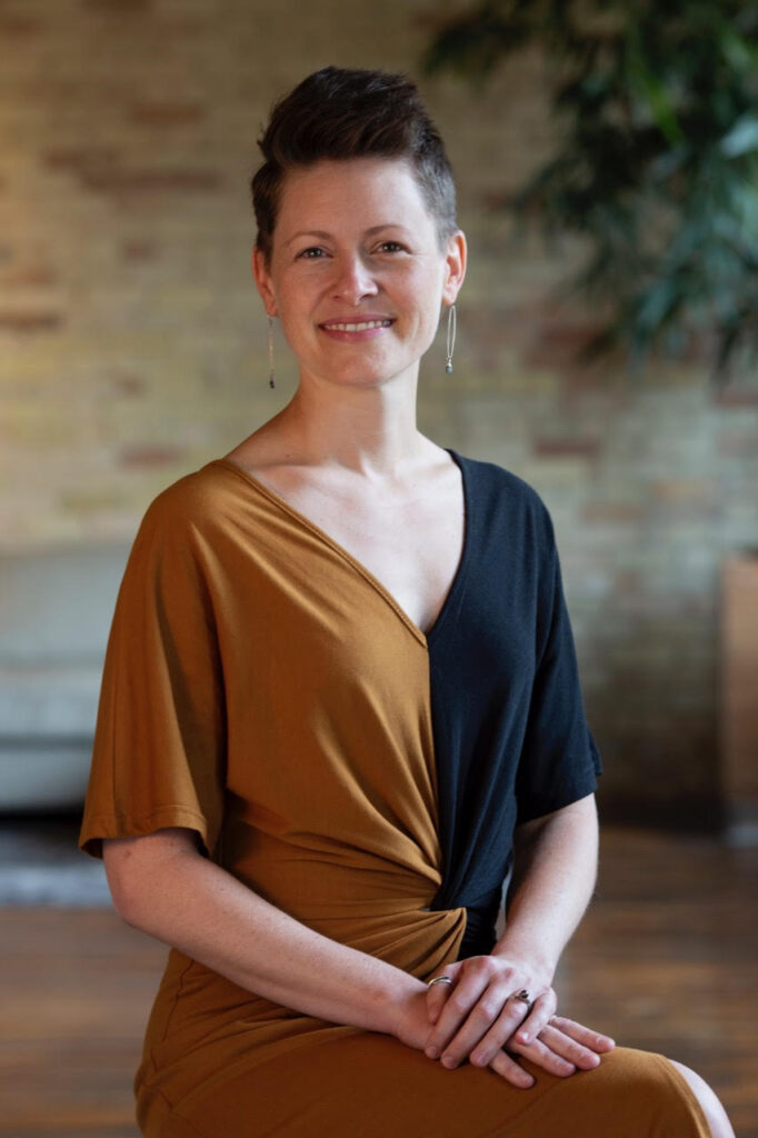A white woman with short dark brown hair, smiling and wearing a dress that is half brow on the left and half black on the right