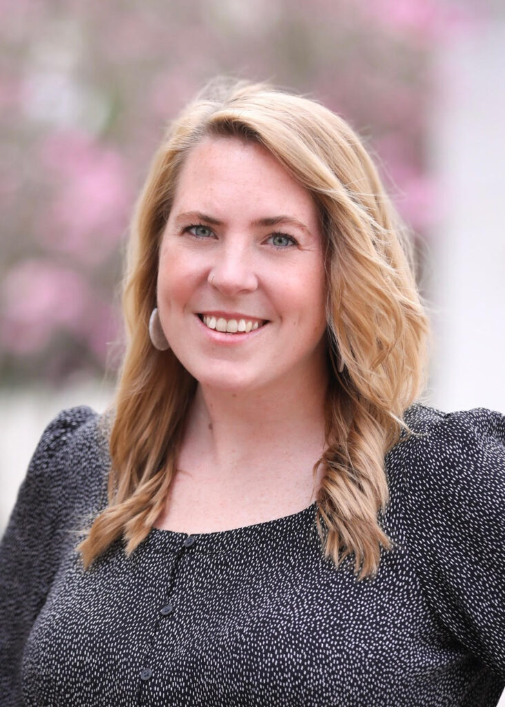 a white woman with shoulder length blonde hair, she smiles and is wearing a black blouse with puff sleeves and tiny white polka dots