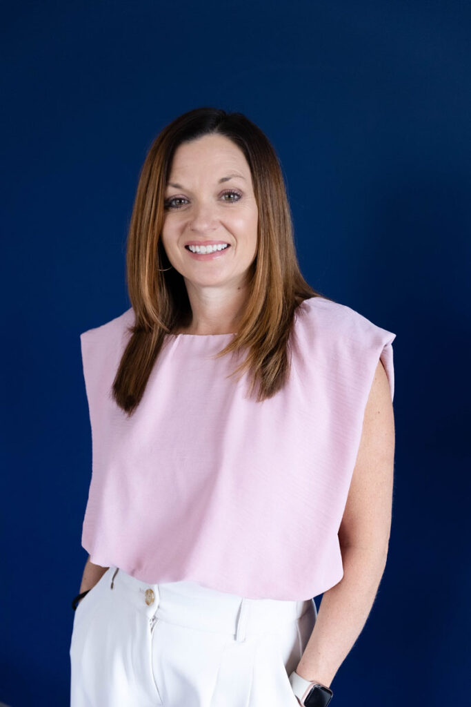 Middle aged white woman smiling with long brown hair with blonde highlights wearing a pink sleeveless shirt.